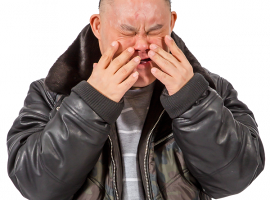 Man covering his face with both hands, wearing a black leather jacket with a fur collar.