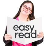 A young woman with Down syndrome, wearing glasses and a pink top, is smiling while holding an open booklet. The booklet has the words "easy read" written in large, bold black letters on a white background.